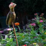 Foto: Galgenvogel auf einem Stab. Blühende Blumen im Hintergrund.